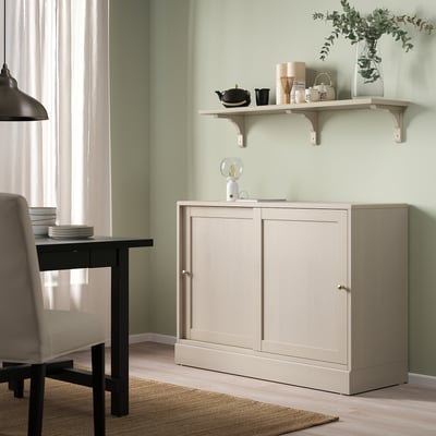 BERGSHULT hylla i beige rymmer tekanna, vaser, växter; ovanför beige skåp, nära svart bord.