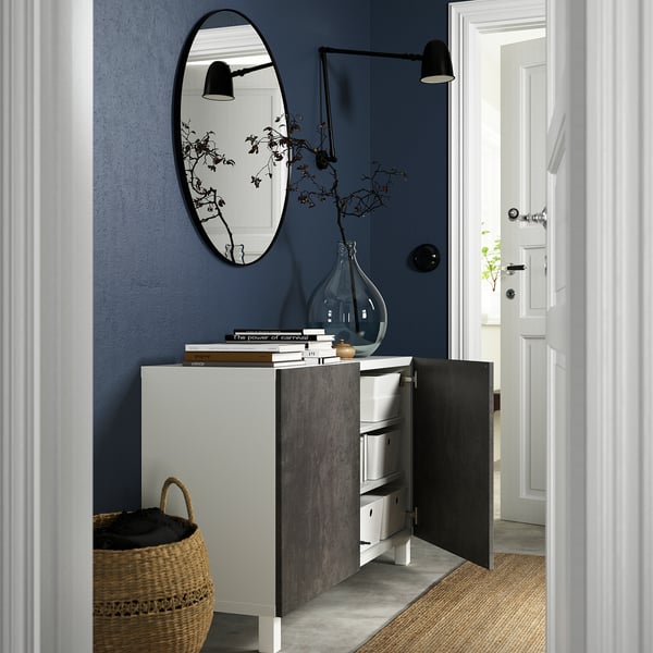 Liten entré med skänken BESTÅ, öppen dörr som avslöjar förvaring. Mörkblå vägg, spegel, svart strömbrytare.