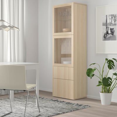 Ljust BESTÅ-skåp i trä med glasdörrar. Hög, smal, passar bra i hörn. Förvarar porslin och glas. Nära vit stol och växt.