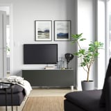 Modernt vardagsrum med TV-bänk BESTÅ, plattskärm, naturfoton, svart lampa och stor grön växt.