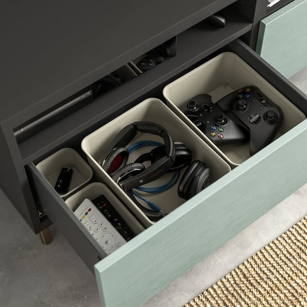 Den öppna lådan visar organiserad teknisk utrustning: spelkontroller, hörlurar, fjärrkontroll och kablar i facken på en ren, grå BESTÅ-enhet.