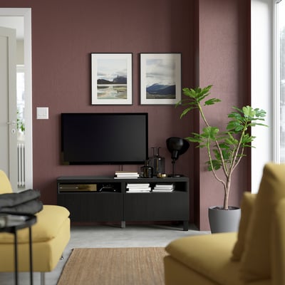 Modernt vardagsrum med rödbruna väggar, gul soffa, tv på svart BESTÅ-stativ och hyllor, växt, inramade foton och mysig matta.