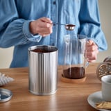 En person skopar upp kaffesump i en metallbehållare på ett bord, med en fransk glaspress och en mugg i närheten.