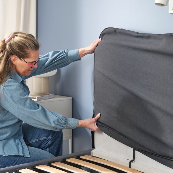 En kvinna ställer i ordning en ottomansk säng från BLÅFJÄLLET och visar förvaringsutrymmet genom att dra i en rem.