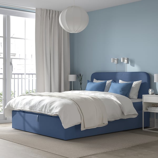 Modernt sovrum med BLÅFJÄLLET blå divan, vita sängkläder, stort fönster, lampa, blå vägg och grå gardiner.