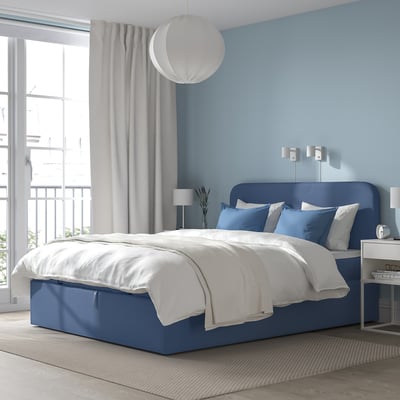 Modern blå ottomansk säng med vita sängkläder, stora fönster, grå gardiner och vägglampor. Mysig och rymlig med en bekväm sänggavel.