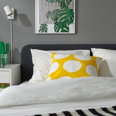 Snyggt sovrum med grå säng, vita kuddar, gul BLÅMUNKAR prickig kudde, grön inredning och grå väggkonst.