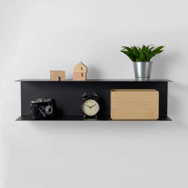 Svart vägghylla med kamera, klocka, trälåda, växt och små dekorativa block. Visar objekt snyggt.