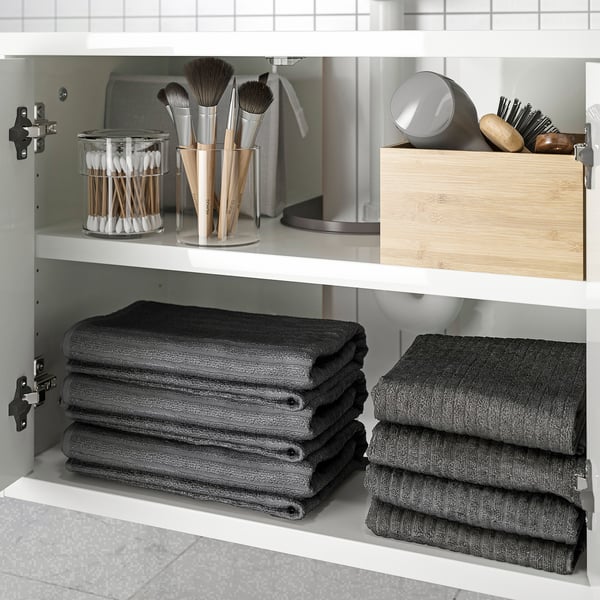 Vitt skåp med handdukar och toalettartiklar: borstar, bomullspinnar i glas, trälåda, grå handdukar vikta.