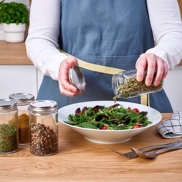 En person kryddar en sallad med örter från en CITRONHAJ kryddburk, andra med kryddor display, på ett köksbord.