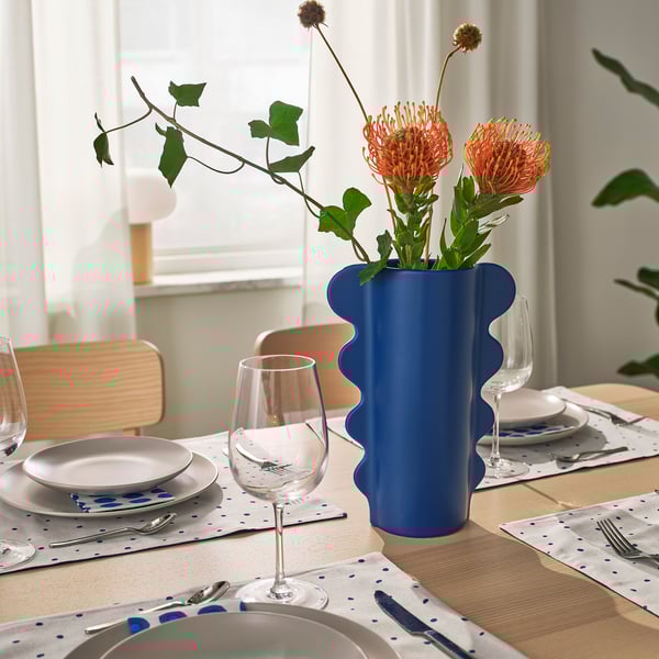 Ett matbord med vita tallrikar, blåprickiga servetter, vinglas och en blå vågig vas med orange blommor.