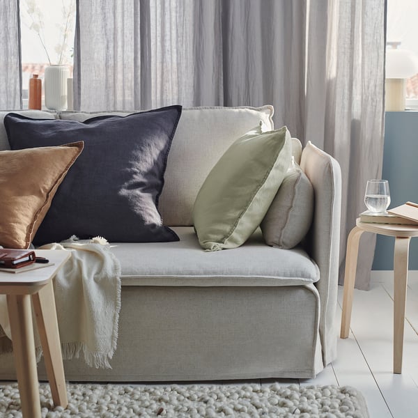 Modernt vardagsrum med ljusgrå soffa, färgglada kuddar, sidobord i trä och skira gardiner.