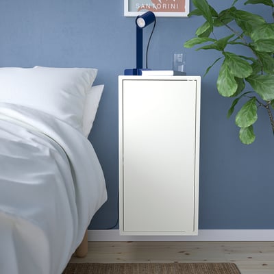 Litet vitt EKET skåp bredvid sängen, blå vägg. Rymmer lampa, växt och böcker. Enkel, modern design.