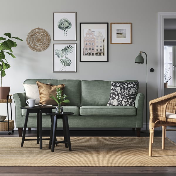 Modernt vardagsrum med soffan EKHOLMA, grön, plysch, 3-sits. Väggkonst, golvlampa, korgstol och matta. Ren, minimalistisk design.