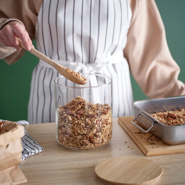 En person skopar granola från en genomskinlig glasburk på ett träbord, perfekt för att förvara och enkelt komma åt torrvaror.