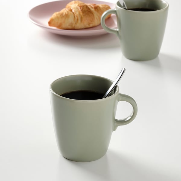 Två gröna muggar med skedar, en fylld med kaffe, bredvid en tallrik med en croissant.