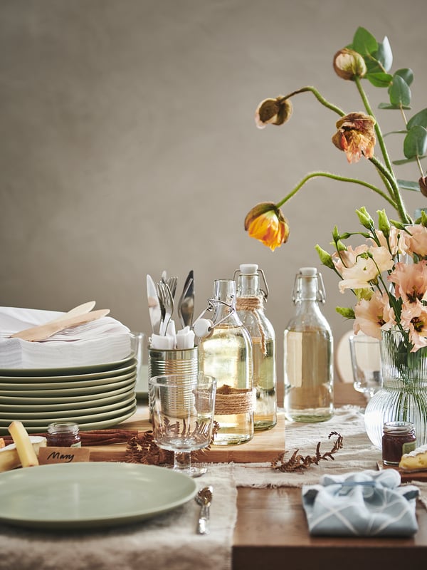 Ett rustikt bordsset med gröna FÄRGKLAR-tallrikar, glasburkar, bestick och blommor.