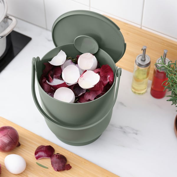 Grön kökssoptunna innehåller organiskt avfall. Locket öppet, skalen synliga. Bänkskiva i marmor och trä med ingredienser i närheten.