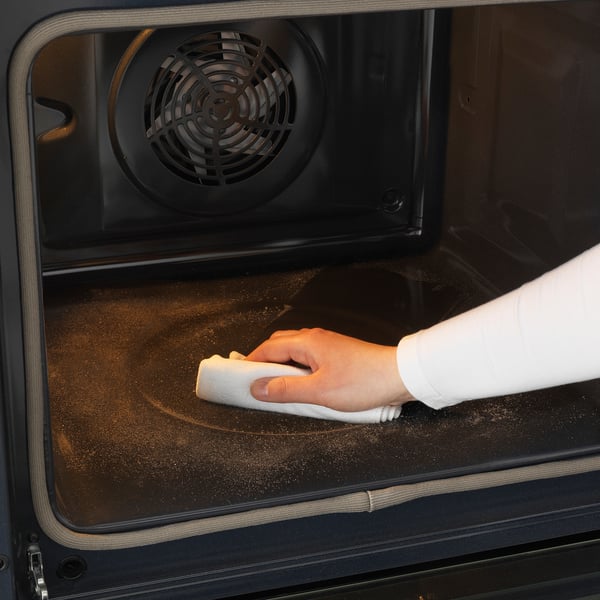 Person som rengör ugnen med trasa. Interiören ser dammig ut. Självrengörande funktion tillgänglig.