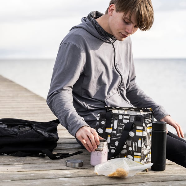 Mannen sitter på bryggan och håller i en grå FLADDRIG matsäck med ett mönster med kaffetema. Bredvid honom står en svart ryggsäck och en flaska.