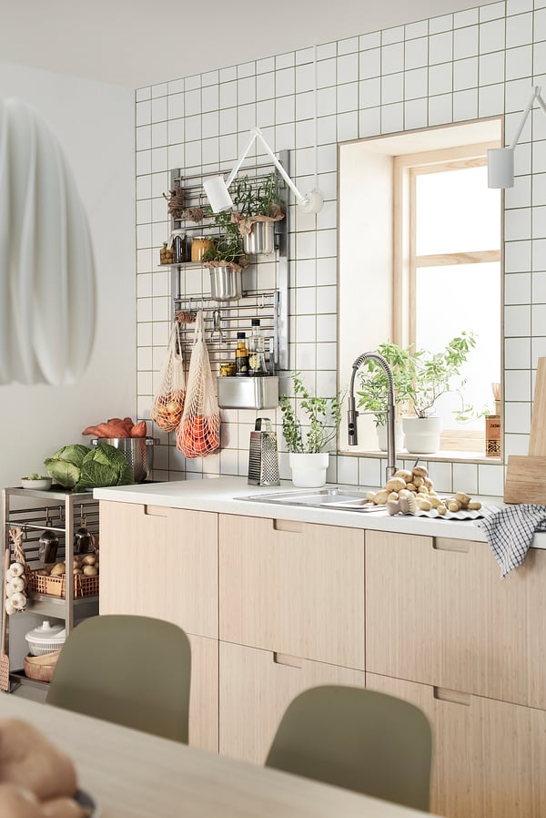 Modernt kök med ljusa träskåp, vitt kakel, gröna stolar, krukväxter och hängande grönsaker.