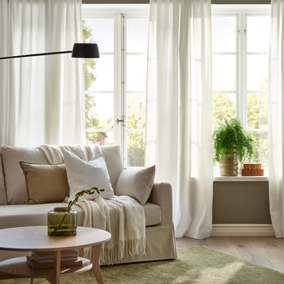Modernt vardagsrum med vita GINSTMOTT gardiner, elegant soffa, mysig pläd och träbord på en grön matta.