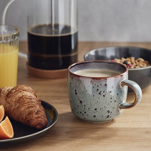Träbord med frukostartiklar: apelsinjuice, croissant, granola och kaffe i en GLADELIG-mugg.