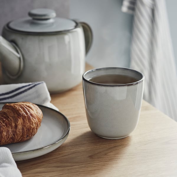 Vit keramisk mugg med mörk glasyr sitter nära tekokare och tallrik med croissant, och visar upp sitt hantverk och enkelhet.