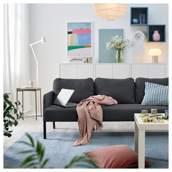 Modernt vardagsrum med svart soffa, grå matta, blå kuddar, bärbar dator, filt, soffbord, lampa, växt och färgglad konst.