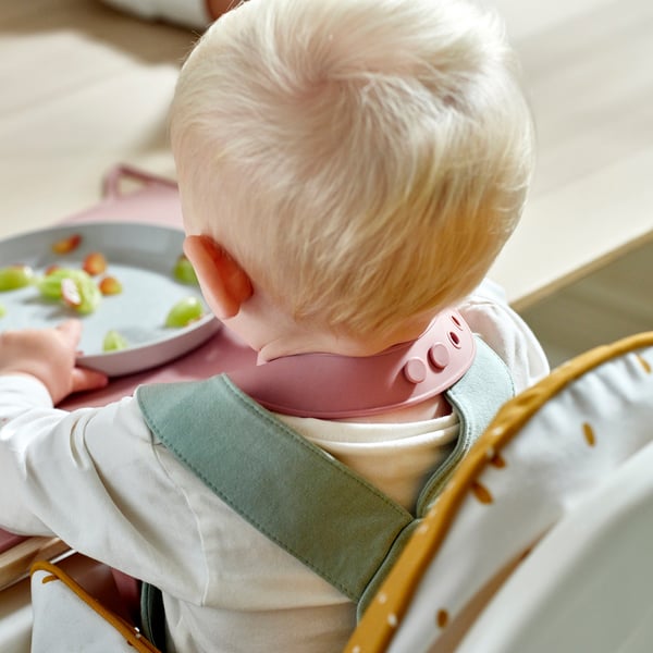Bebis i barnstol som bär silikonhaklapp med en ficka som fångar mat, vilket förhindrar röra på kläderna.