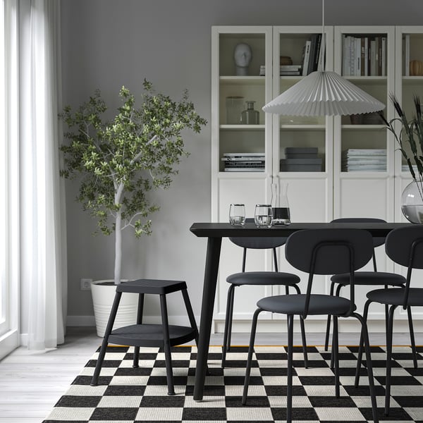 Modern matplats med svarta pallar, stolar, träbord, matta, lampa, hyllor med böcker och en krukväxt.