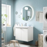 Modernt badrum med svävande vit HAVBÄCK kommod, rund spegel, blått kakel, växter, handdukar, tvättmaskin, träpall.