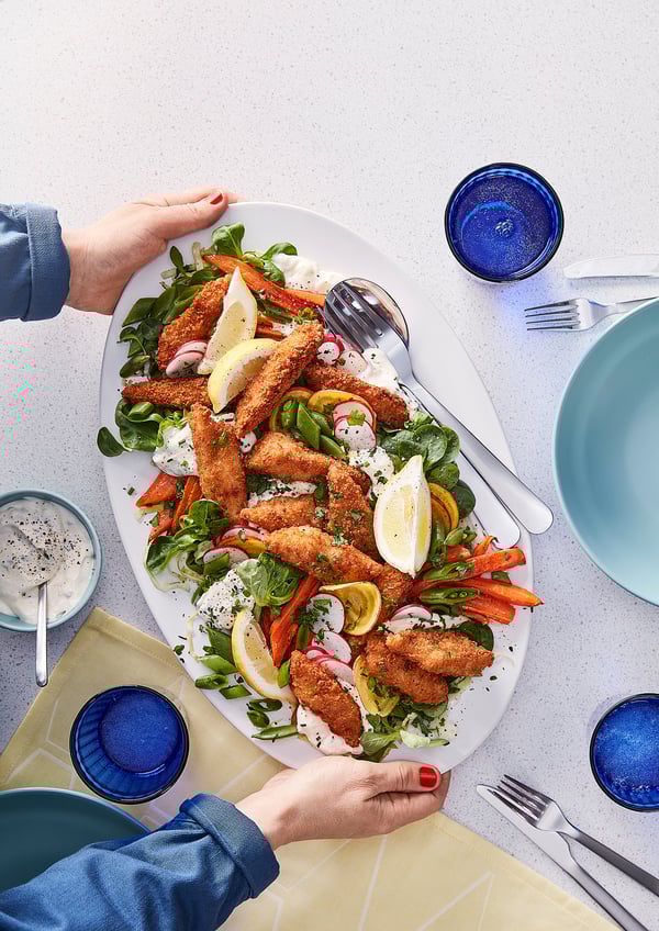 Ett fat med krispiga panerade fiskbitar serverade med sallad. Händer som sträcks ut för att tjäna. Blå dryckeskärl och bestick.