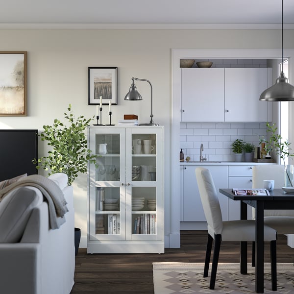 Modernt vardagsrum med vitt furuskåp, glasdörrar. L-format vitt kök, grå lampa, svart matbord med vita stolar.