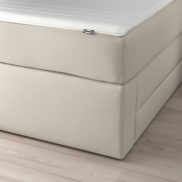 En beige divansäng med lådor för förvaring under madrassen. En IKEA-etikett är synlig. Sängen har en mysig, stoppad design.