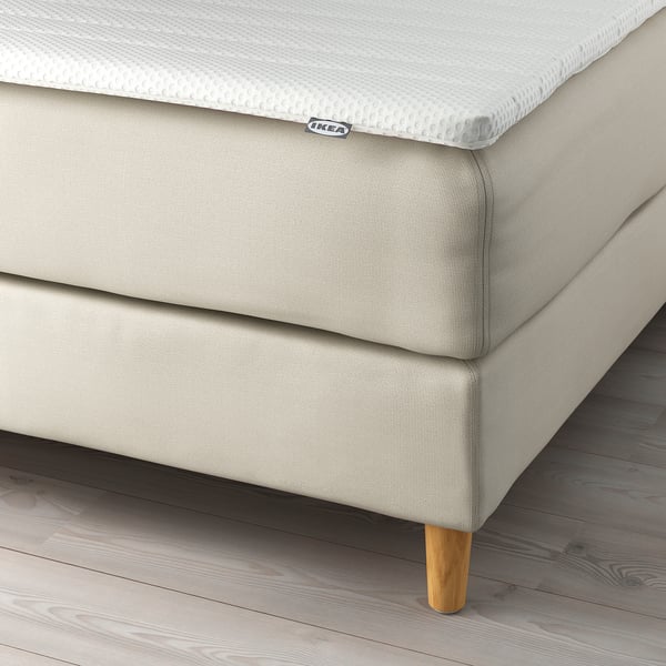 En beige stoppad sängstomme med träben och en vit madrass. IKEA HEMFJÄLLET säng med stödjande huvudgavel för komfort och läsning.