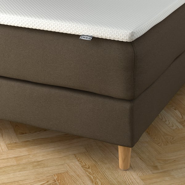 En modern säng med en mjuk, tvättbar sänggavel som erbjuder komfort och enkel skötsel. Ekbenen ger den ett elegant utseende. Taggen visar att det är en IKEA-produkt.