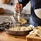 Personen lagar pasta i en stekpanna i rostfritt stål. Såsen hälls över, vilket framhäver pannornas jämna värmefördelning.