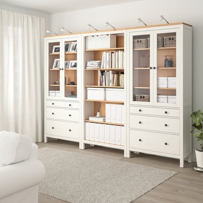 Vit trä HEMNES förvaringskombo med glasdörrar och lådor, visar böcker och inredning, bredvid stora gardiner och soffa.