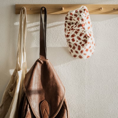 Tre påsar på en klädhängare i trä: brun väska med svart handtag, beige duk, röd och vit fläckig.