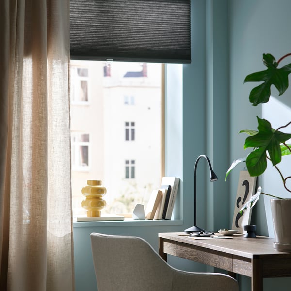 Hemmakontor med blå väggar, grå persienner, skrivbord i trä, lampa, stol, växt och gul vas.