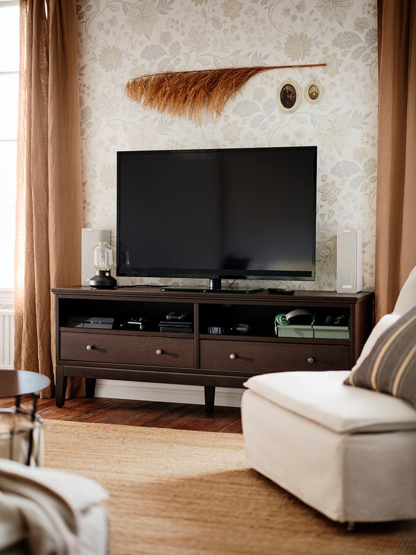 Modernt vardagsrum med IDANÄS tv-bänk, svart plattskärm och fåtölj. Blommiga tapeter, beige gardiner och neutral inredning.