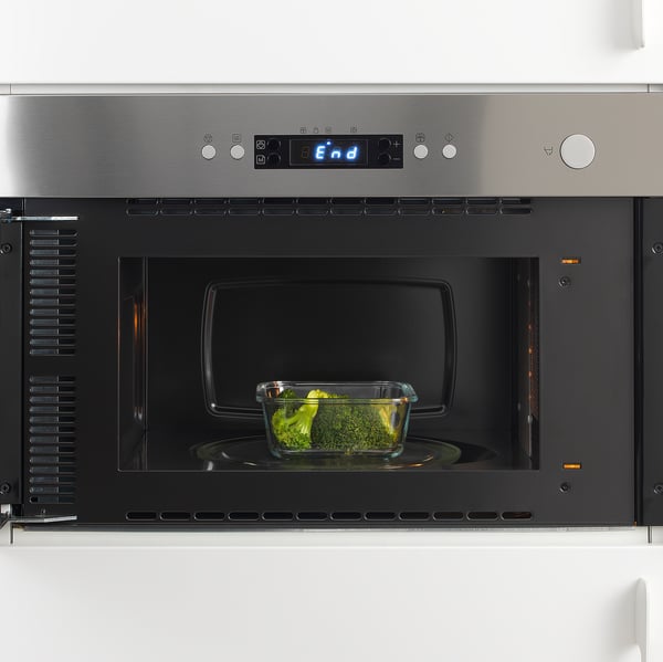 Mikrovågsugn i rostfritt stål med glasbehållare med broccoli inuti, som visar dess funktion att värma eller tina mat.