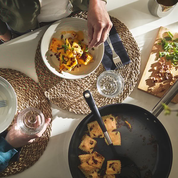 En person som serverar tillagad mat från en stekpanna på en tallrik. Stekpannan står på ett bord med bordstabletter och glas.