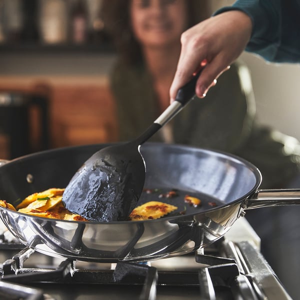En person lagar mat i en stor, non-stick IKEA 365+ stekpanna på en spis och rör om gula omelettbitar med en spatel. Pannan har två handtag för enkel lyftning.