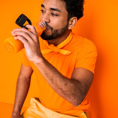 Man i orange skjorta som dricker från klar flaska med svart lock och rem, mot orange bakgrund.