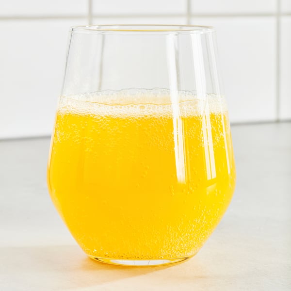 Mugg i klart glas fylld med bubblig, orangefärgad ISKUB-dryck, placerad på en yta. Nyckelingredienser: apelsinarom, sötningsmedel, lågt socker.