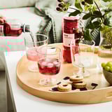 Person som häller rosa dryck i ett glas på en träbricka som innehåller kakor, druvor och en flaska märkt ISKUB lingonsmak.