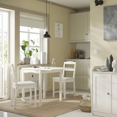 Mysig kökshörna med JUSTINA vita stolar, beige matta, grön växt och skåp i ljust trä.