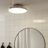 En modern taklampa, som avger diffust ljus, perfekt för badrum, monterad på en vit kaklad vägg ovanför en rund spegel med krokar.